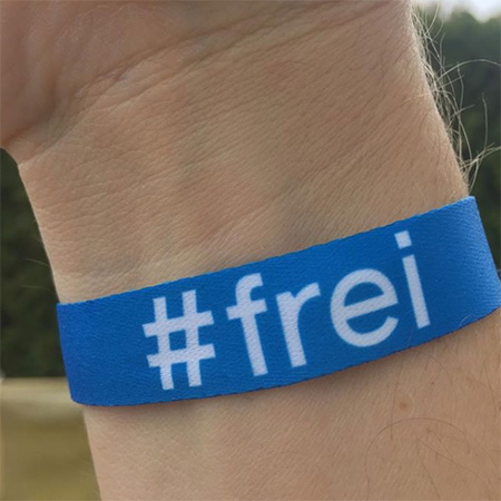 Das Foto zeigt einen Arm mit blauem #frei Band, der ins Bild ragt. Im Hintergrund Natur.