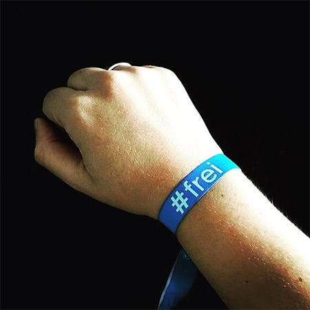 Das Foto zeigt eine Hand mit blauem #frei Band, die ins Bild ragt. Der Hintergrund ist schwarz.