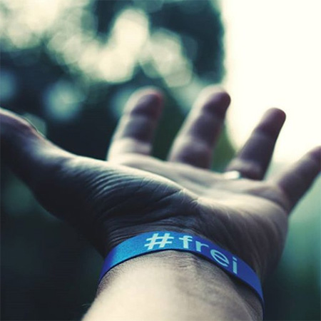 Das Foto zeigt eine ausgestreckte Hand mit blauem #frei Band, die ins Bild ragt. Im Hintergrund erkennt man verschwommen einen Baum.
