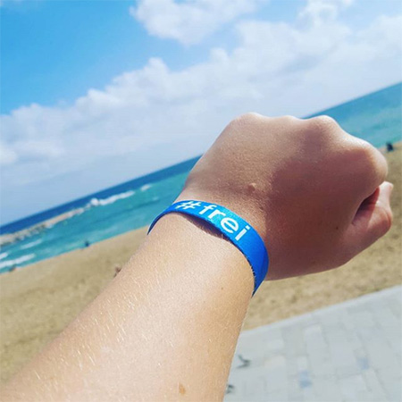 Das Foto zeigt eine ausgestreckte Hand mit blauem #frei Band, die ins Bild ragt. Im Hintergrund erkennt man Strand, Meer und Himmel.