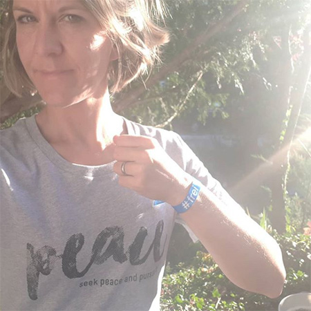 Das Foto zeigt den Oberkörper einer blonden Frau, die ihren Arm mit blauem #frei Band in die Kamera hält. Das Bild ist etwas überbelichtet von den Sonnenstrahlen. Sie steht in der Natur.