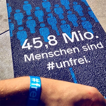 Das Foto zeigt ein Banner mit der Aufschrift „45,8 Mio. Menschen sind #unfrei.“ Davor eine Hand mit blauem #frei Band, die ins Bild ragt.