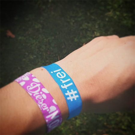 Das Foto zeigt eine ausgestreckte Hand mit einem rosaroten Band und einem blauen #frei Band, die ins Bild ragt.