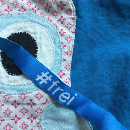 Das Foto zeigt ein blaues #frei Band, auf einer gemusterten Stoffdecke.