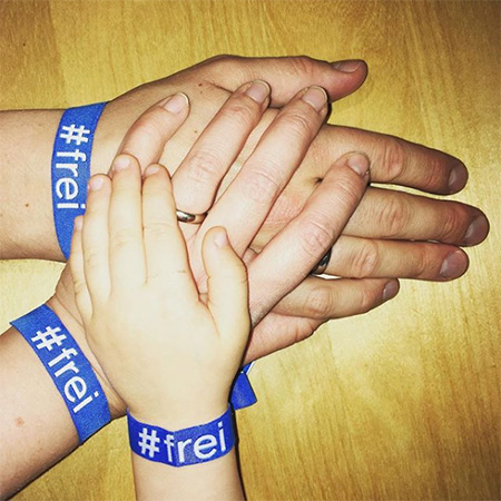 Das Foto zeigt drei ausgestreckte Hände mit blauem #frei Band, die ins Bild ragen. Die Hände liegen aufeinander. Zuoberst eine Kinderhand.
