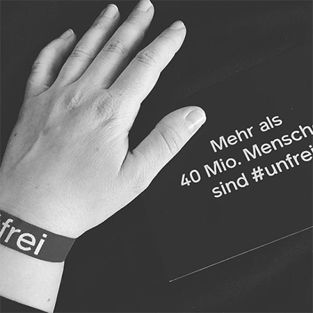 Das Foto zeigt einen Arm mit dem blauen #frei Band, der in die Kamera ragt.  Im Hintergrund eine Karte, auf der steht „Mehr als 40 Mio. Menschen sind #unfrei“. Das Bild ist schwarz/weiß