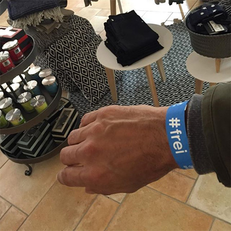 Das Foto zeigt eine ausgestreckte Männerhand mit blauem #frei Band, die ins Bild ragt. Im Hintergrund mehrere Tische.