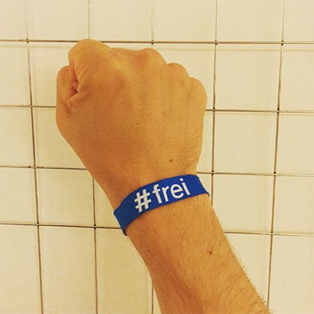 Das Foto zeigt eine ausgestreckte Männerhand mit blauem #frei Band, die ins Bild ragt. Im Hintergrund weiße Fließen.