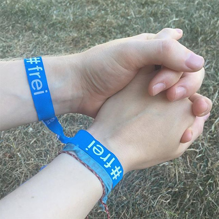 Das Foto zeigt zwei Hände, die in Bild ragen und sich in der Bildmitte falten - mit blauem #frei Band. Im Hintergrund Wiese.