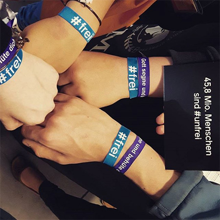 Das Foto zeigt vier Arme mit blauem #frei Band, die aus verschiedenen Richtungen in die Kamera ragen, nebst einer Karte, auf der steht „Mehr als 40 Mio. Menschen sind #unfrei“.