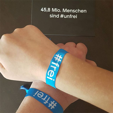 Das Foto zeigt zwei gekreuzte Arme mit blauem #frei Band, nebst einer Karte, auf der steht „Mehr als 40 Mio. Menschen sind #unfrei“.