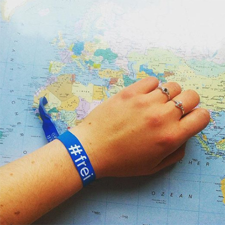 Das Foto zeigt eine ausgestreckte Frauenhand mit blauem #frei Band, die ins Bild ragt. Im Hintergrund ein Globus.