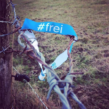 Das Foto zeigt ein blaues #frei Band, das an einem Stacheldrahtzaun hängt.