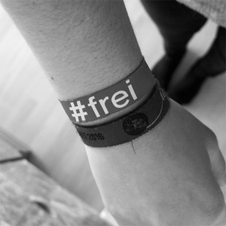 Das Foto zeigt einen ausgestreckten Arm mit einem #frei Band, der von oben ins Bild gehalten wird. Das Bild ist schwarz/weiß.