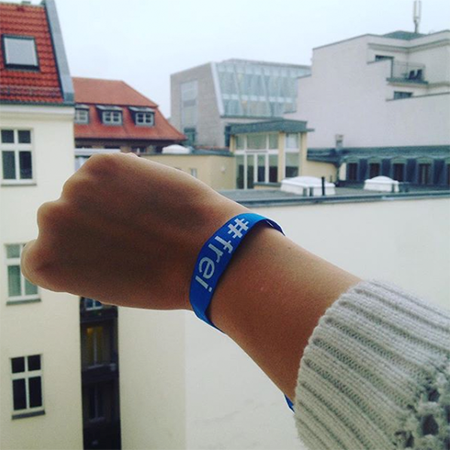 Das Foto zeigt einen Arm mit #frei Band am Handgelenk. Im Hintergrund sind Häuser zu sehen.