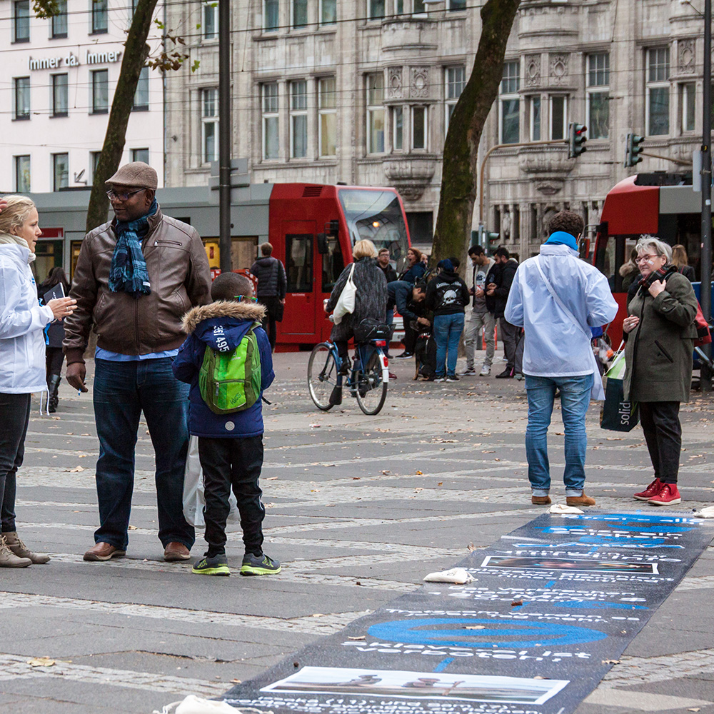 Das Bild zeigt mehrere Menschen auf der Straße in einer Fußgängerzone. Auf dem Boden eine blaue Malerei.