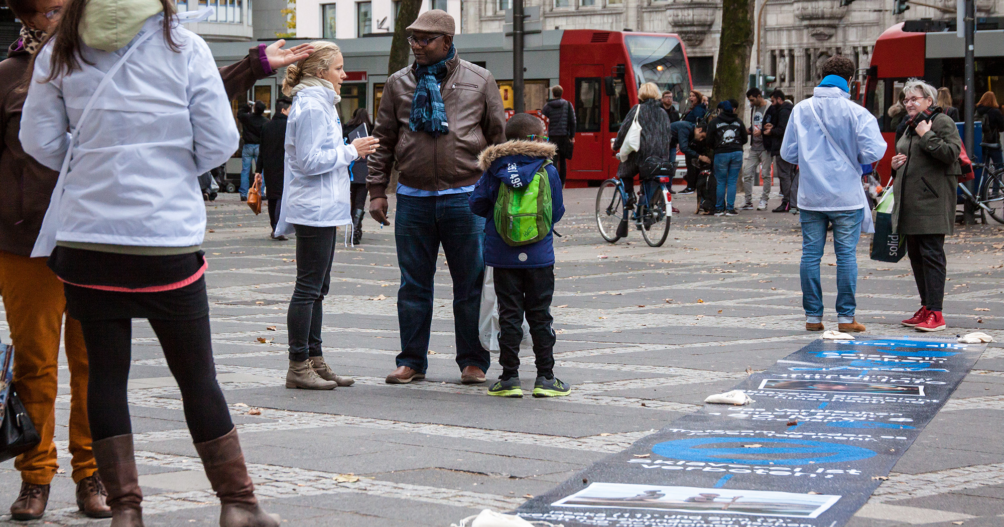 Das Bild zeigt mehrere Menschen auf der Straße in einer Fußgängerzone. Auf dem Boden eine blaue Malerei.