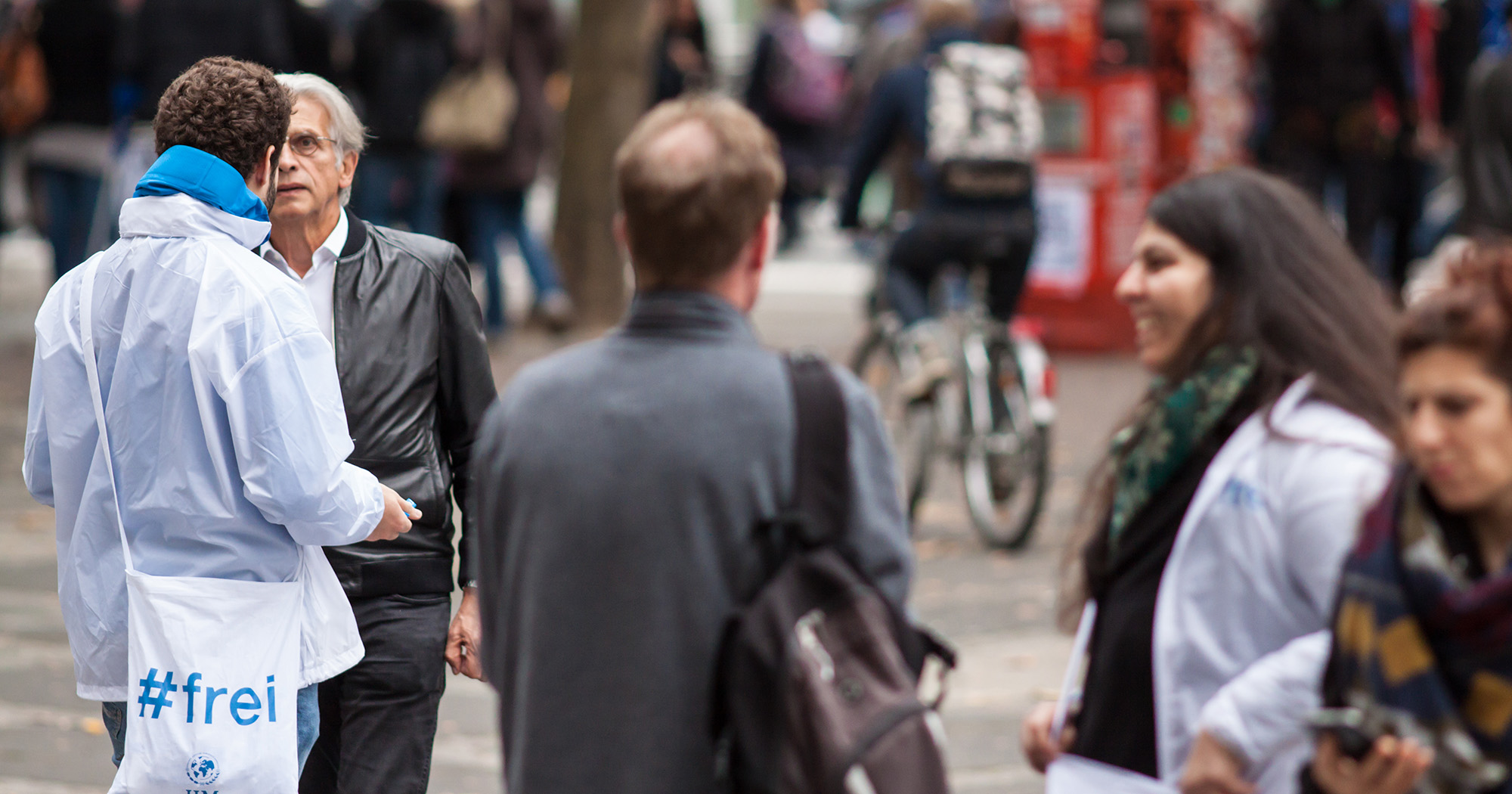 Das Bild zeigt einen Mann von hinten auf der Straße mit einer Tüte um die Schulter auf der „#frei“ steht. Er redet mit einem anderen Mann im Anzug.