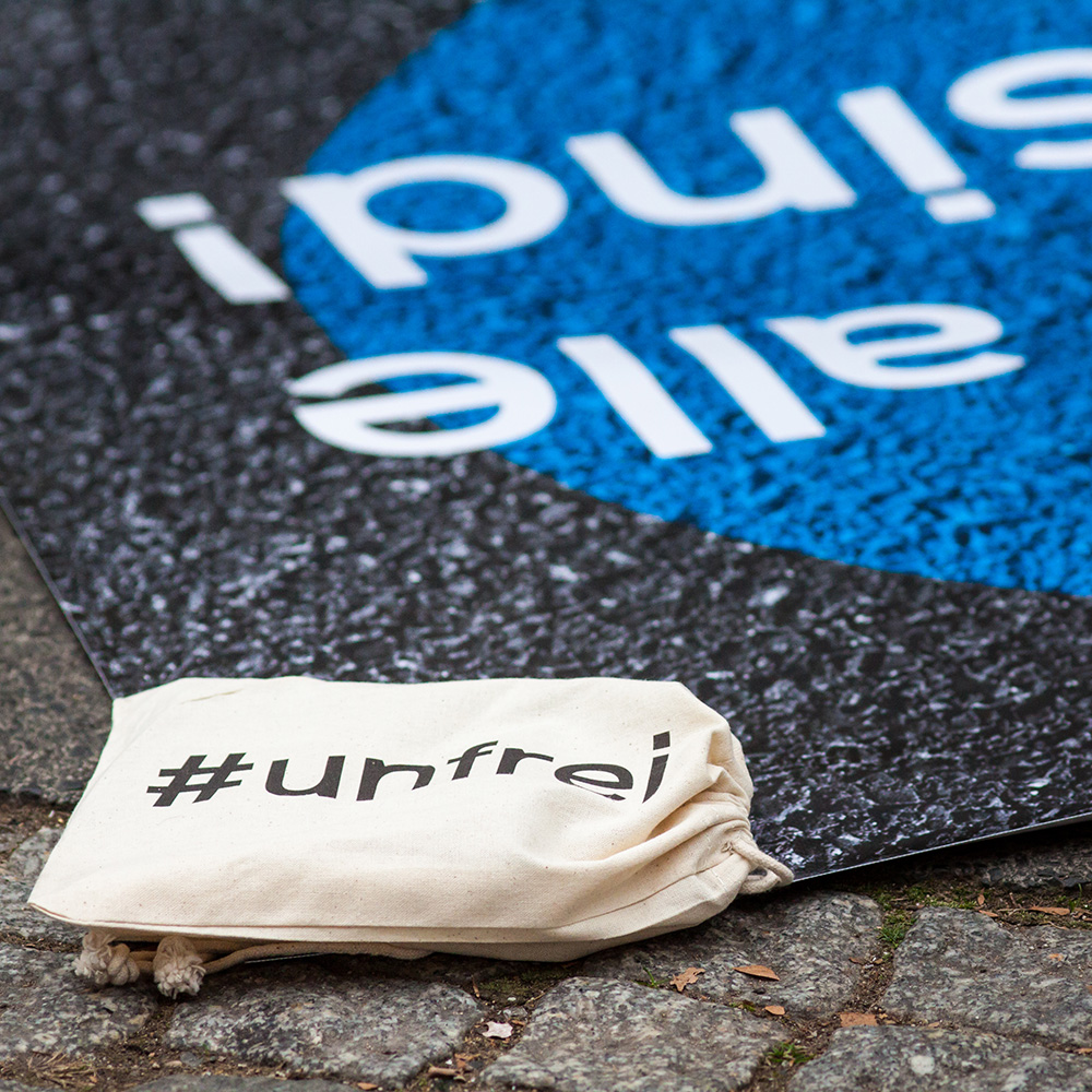 Das Bild zeigt den Boden einer Straße mit blauer Malerei und einem kleinen Jutebeutel, auf dem „#frei“ steht.