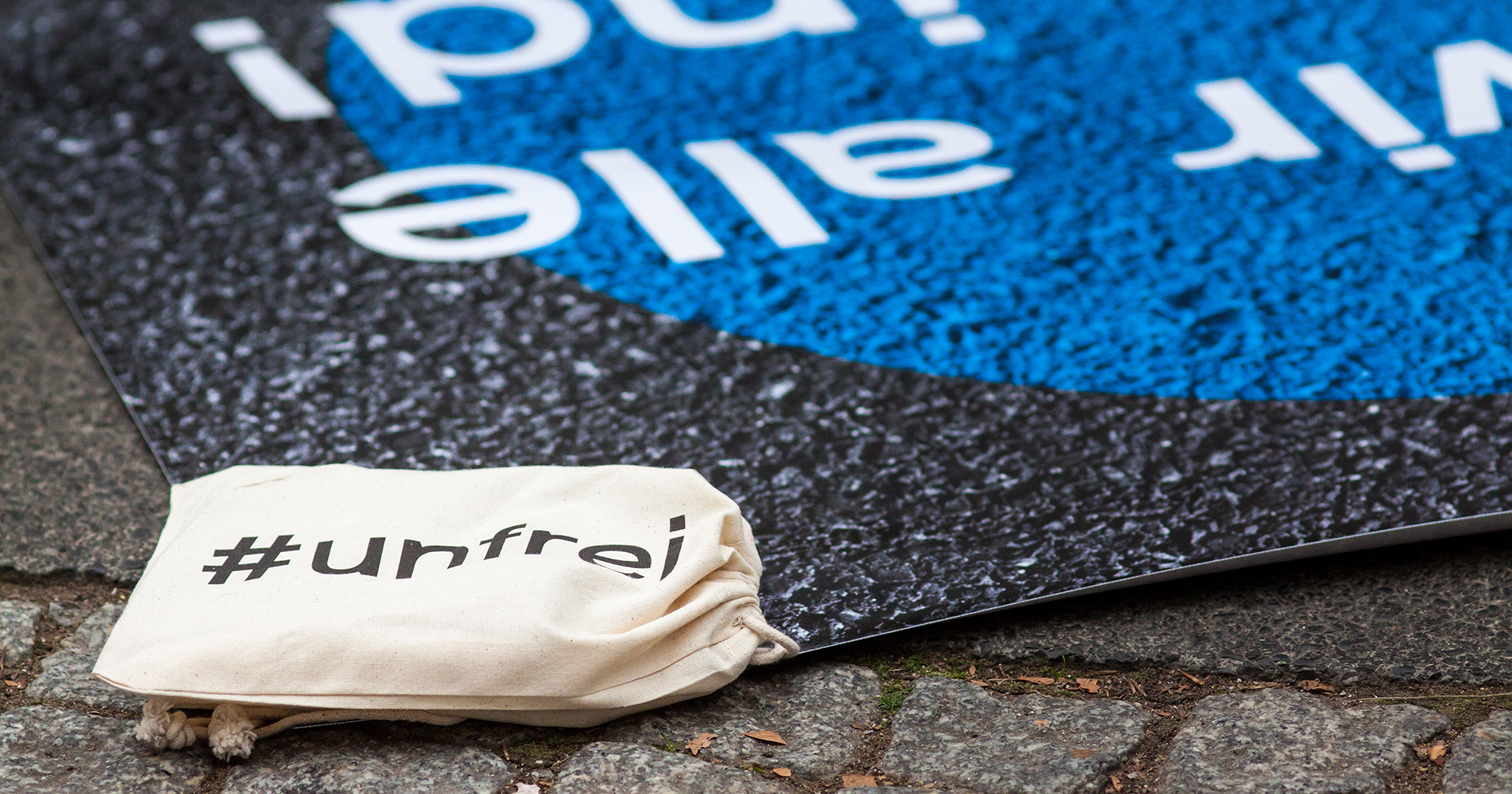 Das Bild zeigt den Boden einer Straße mit blauer Malerei und einem kleinen Jutebeutel, auf dem „#frei“ steht.