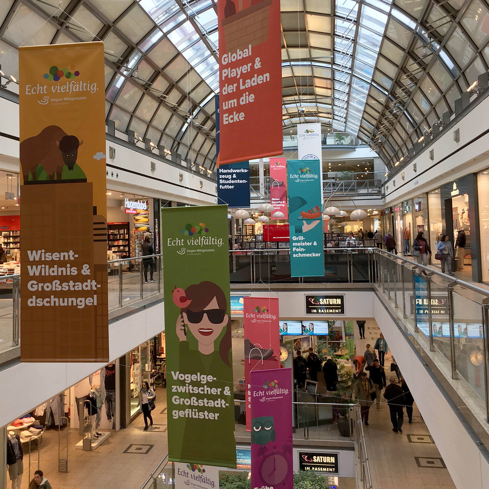 Das Foto zeigt eine Shoppingmall von innen. Darin hängen die SiWi-Motive als Banner von der Decke.