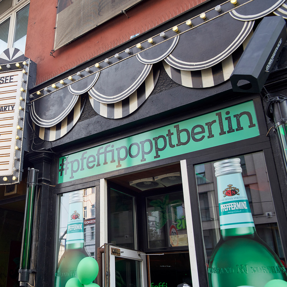Das Foto zeigt den Eingangsbereich einer Bar von außen. Über der Tür steht „#pfeffipopptberlin“, seitlich neben der Tür sind zwei große Pfeffi Flaschen aufgedruckt.