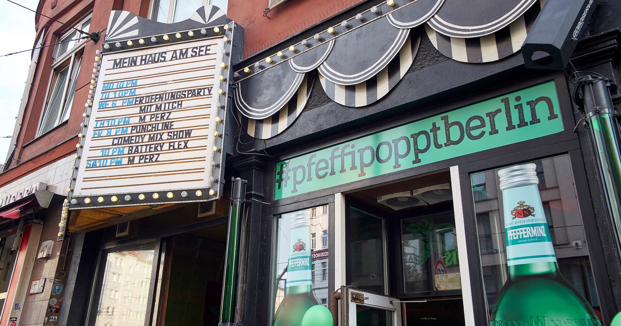 Das Foto zeigt den Eingangsbereich einer Bar von außen. Über der Tür steht „#pfeffipopptberlin“, seitlich neben der Tür sind zwei große Pfeffi Flaschen aufgedruckt.