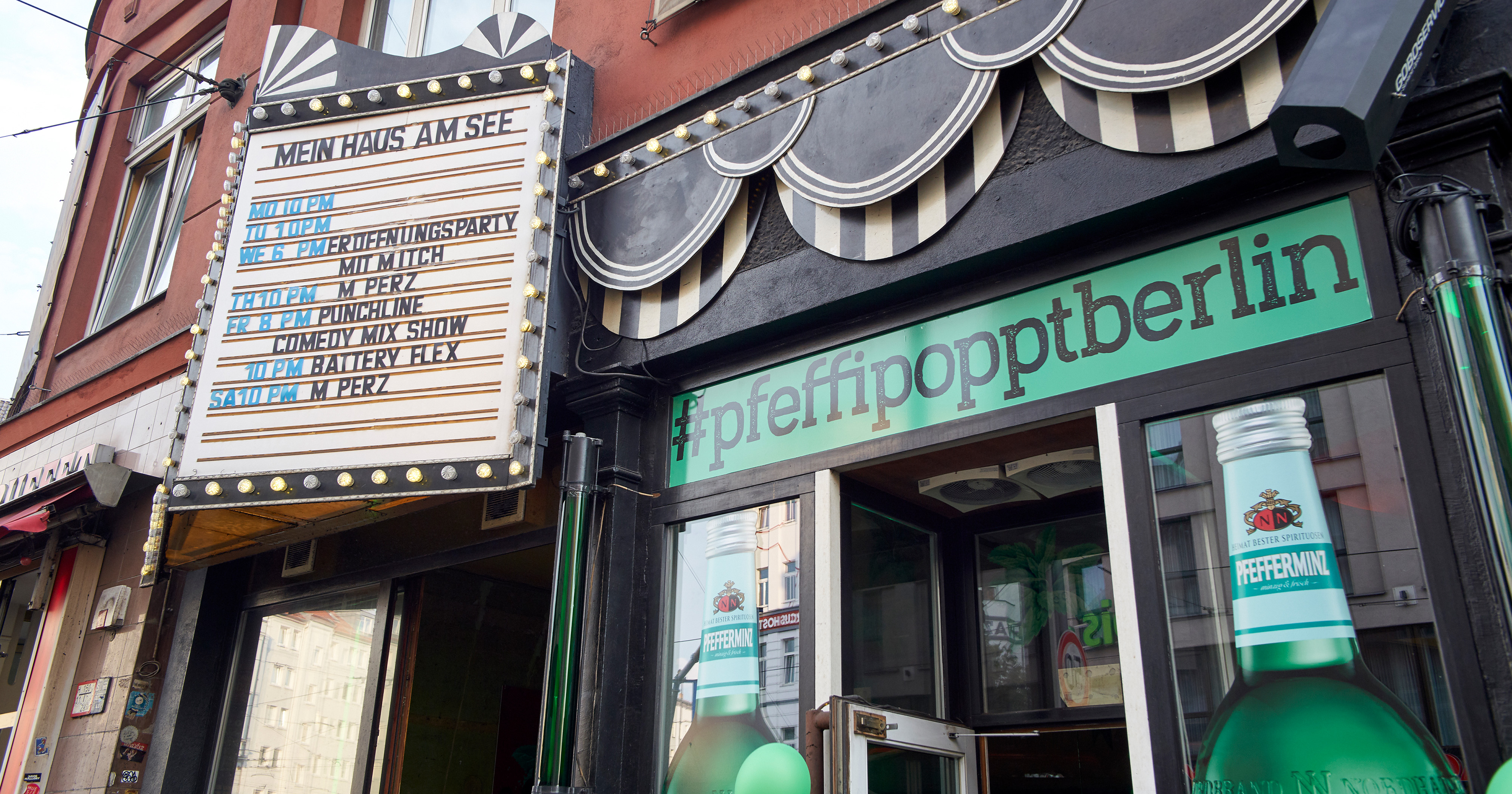 Das Foto zeigt den Eingangsbereich einer Bar von außen. Über der Tür steht „#pfeffipopptberlin“, seitlich neben der Tür sind zwei große Pfeffi Flaschen aufgedruckt.