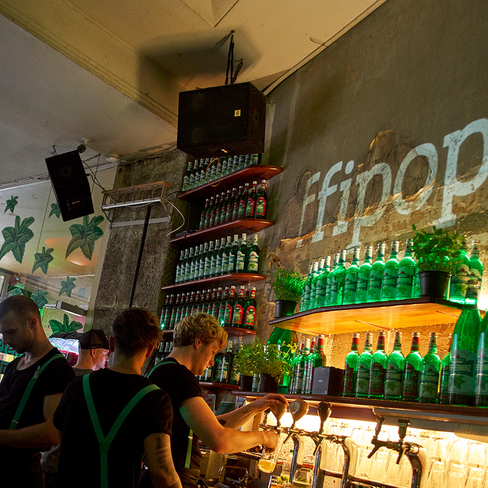 Das Foto zeigt die Theke in einer Bar mit einem Flaschenregal voll mit Pfeffi Flaschen. Hinter der Theke mehrere Barkeeper mit grünen Hosenträgern.