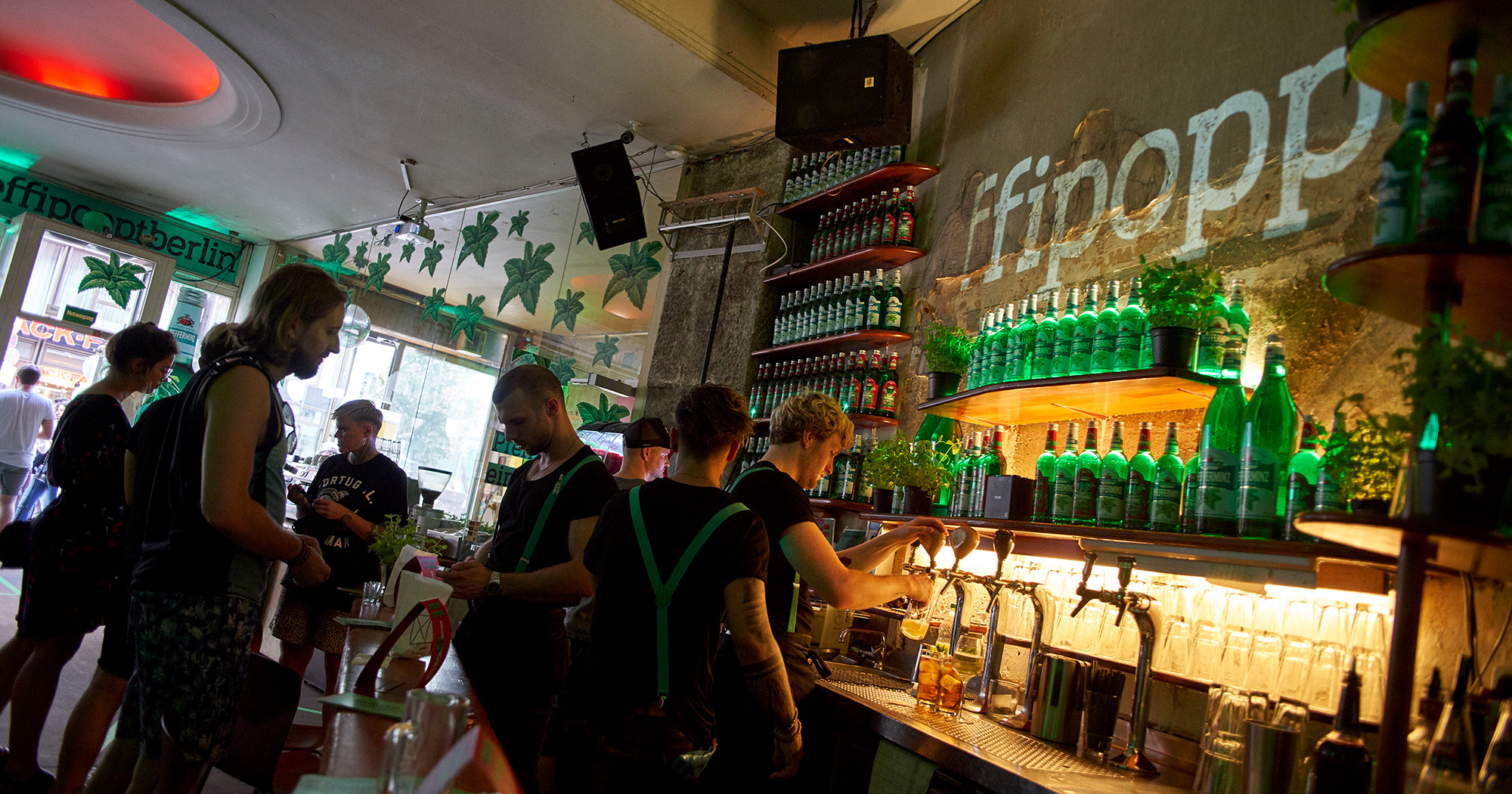 Das Foto zeigt die Theke in einer Bar mit einem Flaschenregal voll mit Pfeffi Flaschen. Hinter der Theke mehrere Barkeeper mit grünen Hosenträgern.