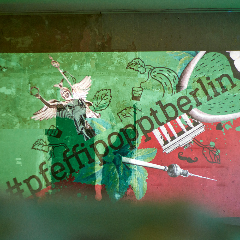 Das Foto zeigt das Logo von „#pfeffipopptberlin“ mit Illustrationen der Siegessäule, des Brandenburger Tors und des Fernsehturms sowie einem Mund mit grüner Zunge auf rot/grünem Hintergrund.