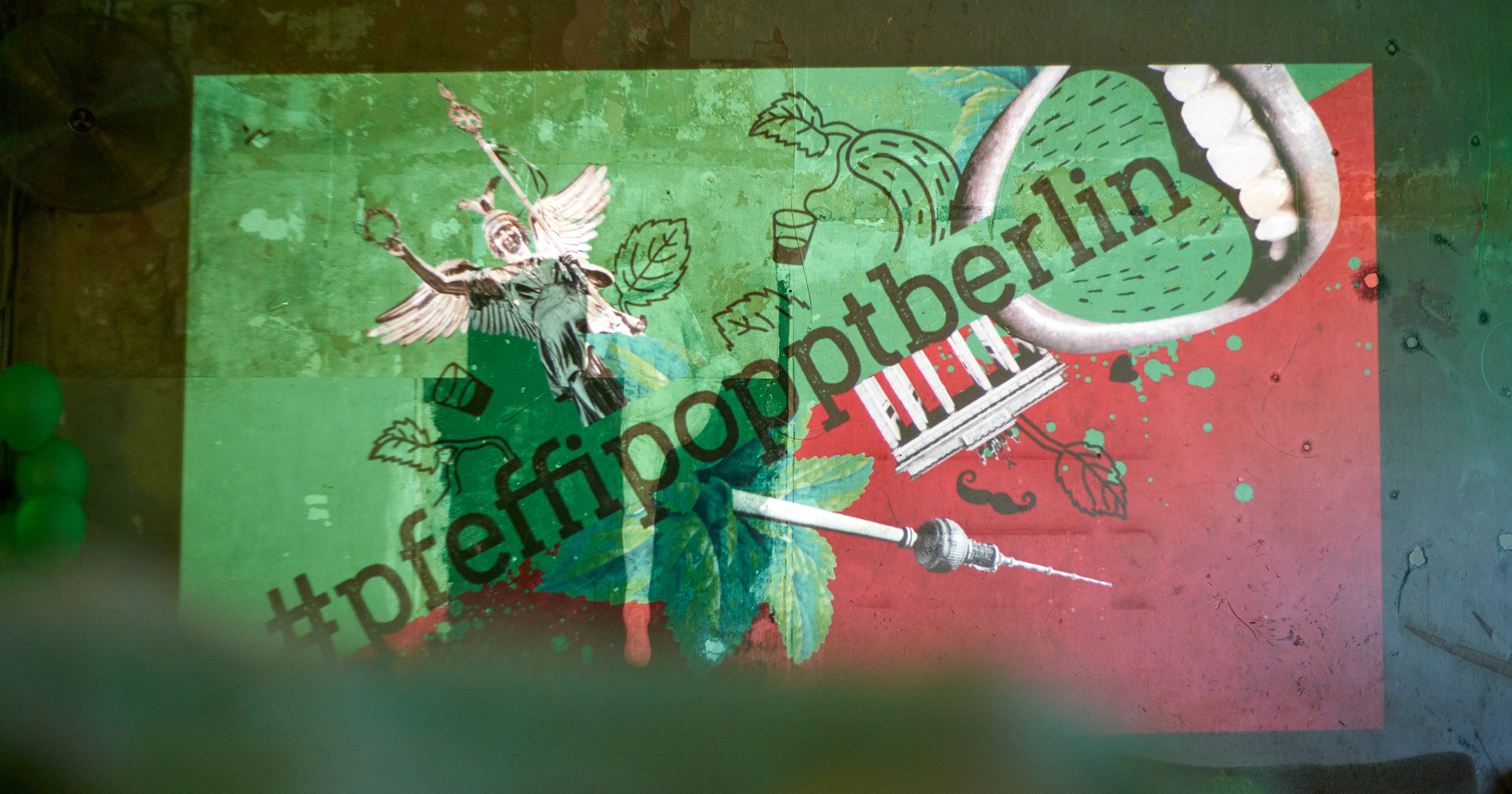 Das Foto zeigt das Logo von „#pfeffipopptberlin“ mit Illustrationen der Siegessäule, des Brandenburger Tors und des Fernsehturms sowie einem Mund mit grüner Zunge auf rot/grünem Hintergrund.