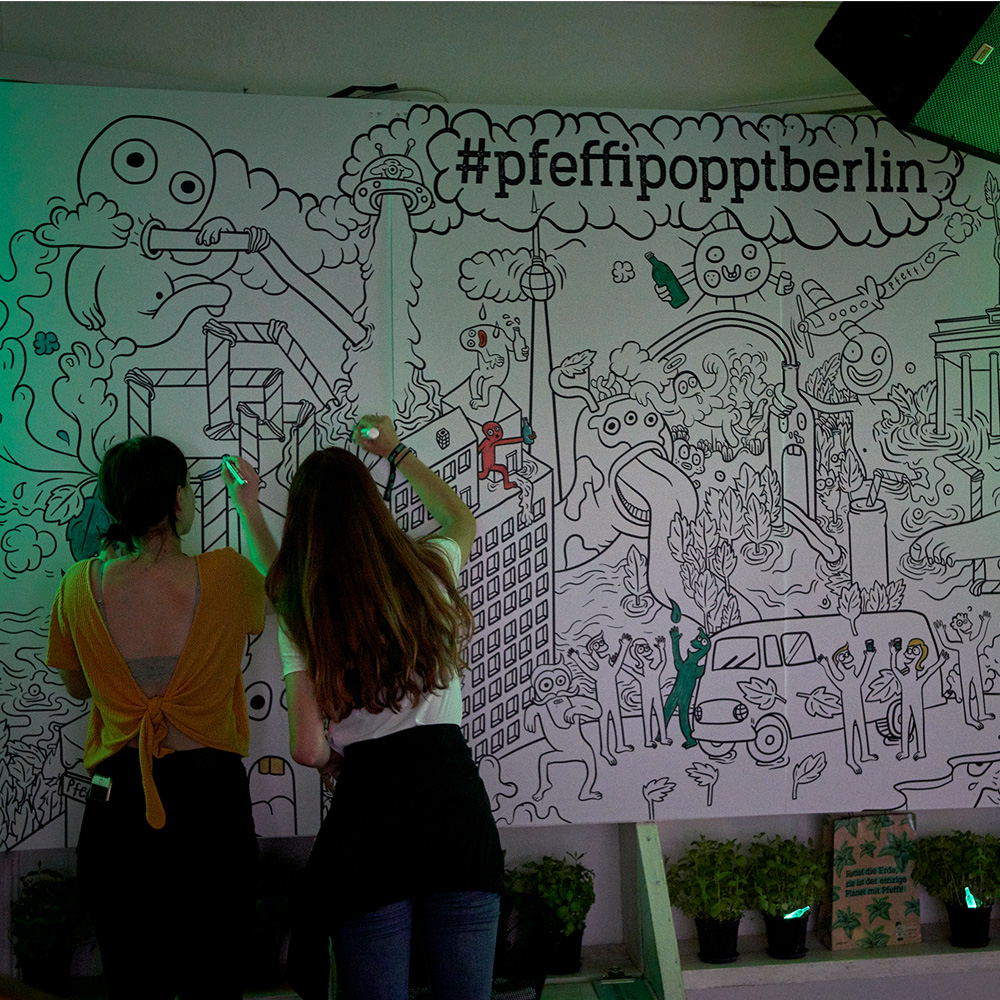Das Foto zeigt die Mandala-Wall im „#pfeffipopptberlin“ Popupstore. Man sieht unter anderem lustige Phantasiewesen sowie Illustrationen des Brandenburger Tors und des Fernsehturms. Davor Menschen, die das Bild ausmalen.