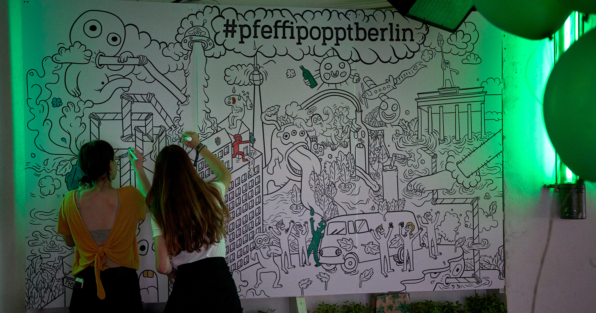 Das Foto zeigt die Mandala-Wall im „#pfeffipopptberlin“ Popupstore. Man sieht unter anderem lustige Phantasiewesen sowie Illustrationen des Brandenburger Tors und des Fernsehturms. Davor Menschen, die das Bild ausmalen.