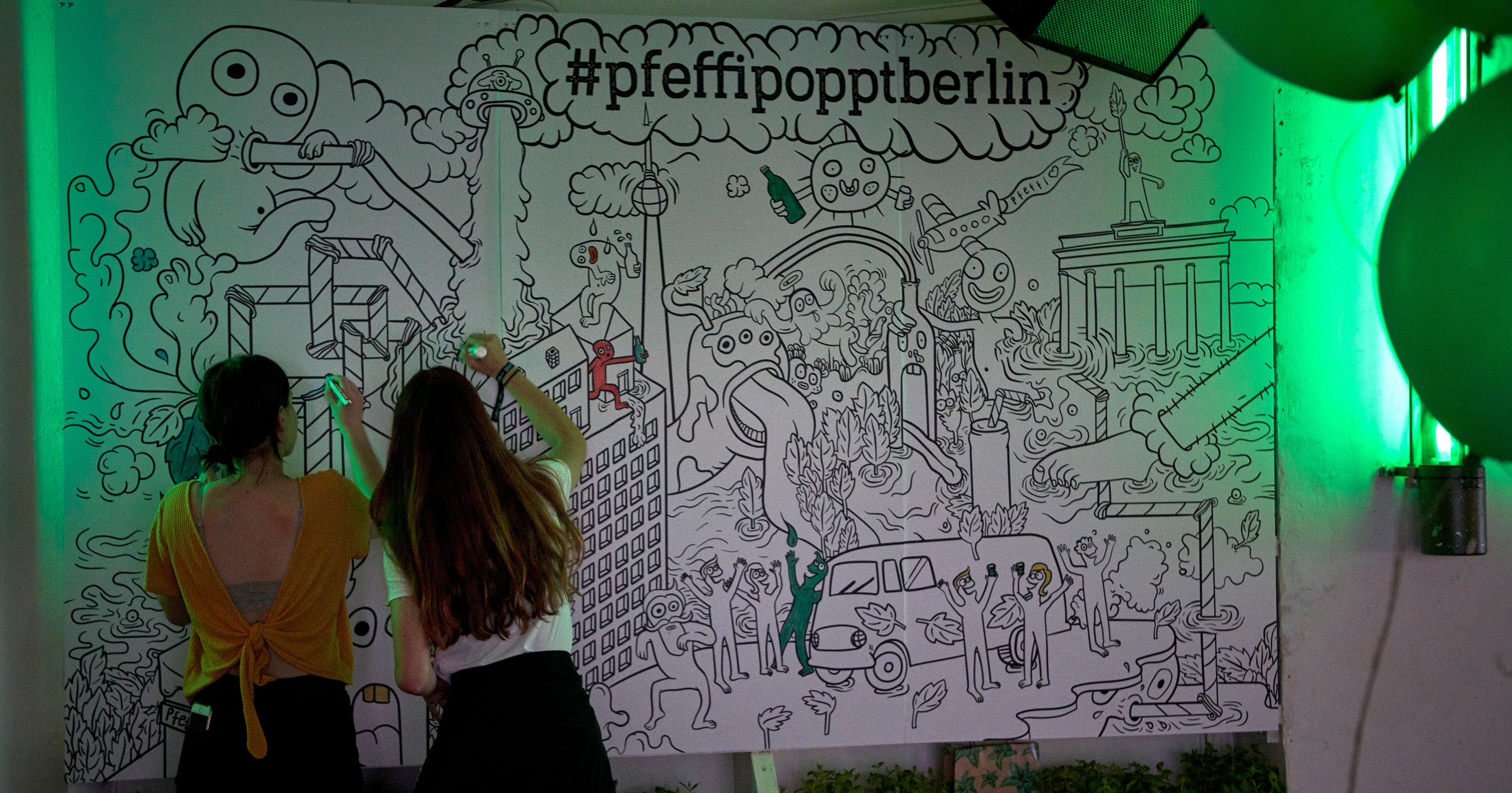 Das Foto zeigt die Mandala-Wall im „#pfeffipopptberlin“ Popupstore. Man sieht unter anderem lustige Phantasiewesen sowie Illustrationen des Brandenburger Tors und des Fernsehturms. Davor Menschen, die das Bild ausmalen.