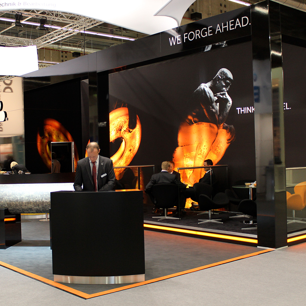 Das Foto zeigt den Infobereich des Messestands von RKind. Im Hintergrund Menschen auf Sesseln, die sich unterhalten. Hinter ihnen eine Wand, auf der Denker von RKind abgebildet ist. 