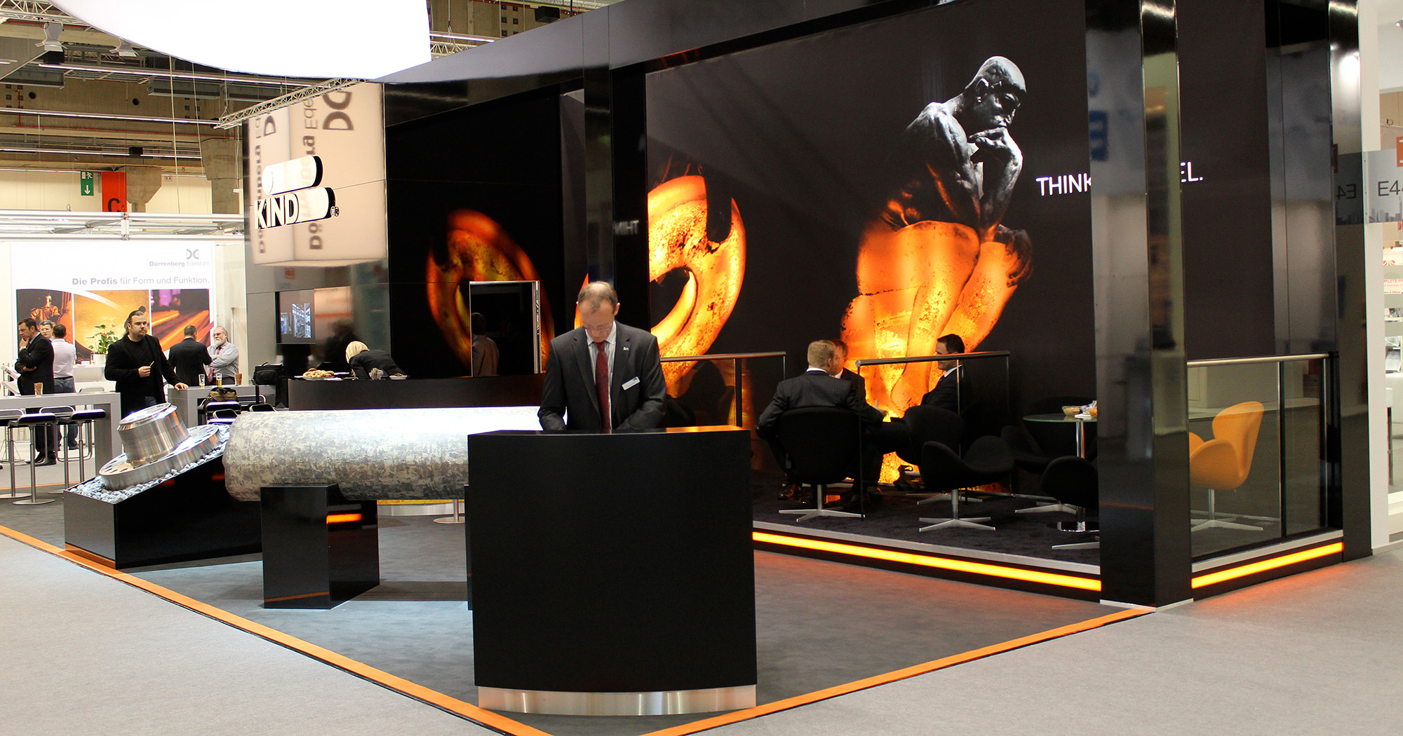 Das Foto zeigt den Infobereich des Messestands von RKind. Im Hintergrund Menschen auf Sesseln, die sich unterhalten. Hinter ihnen eine Wand, auf der Denker von RKind abgebildet ist. 